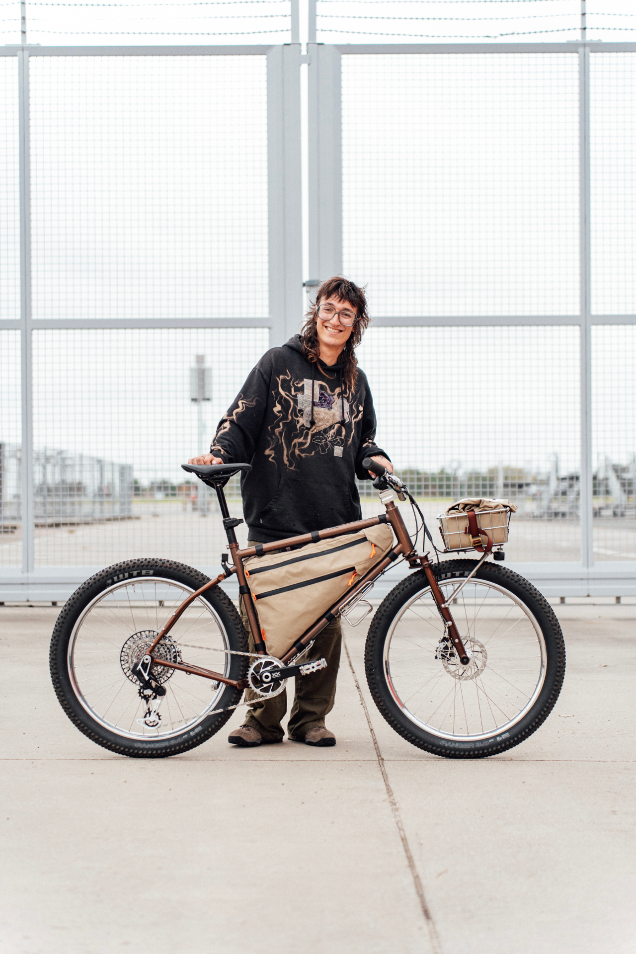 Framebuilder smiling beside their custom brown steel bike, with their full head visible in frame