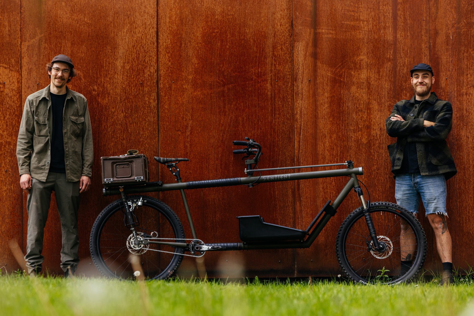 Two builders standing beside a Sentient Apocalypse cargo bike in front of a corten steel wall